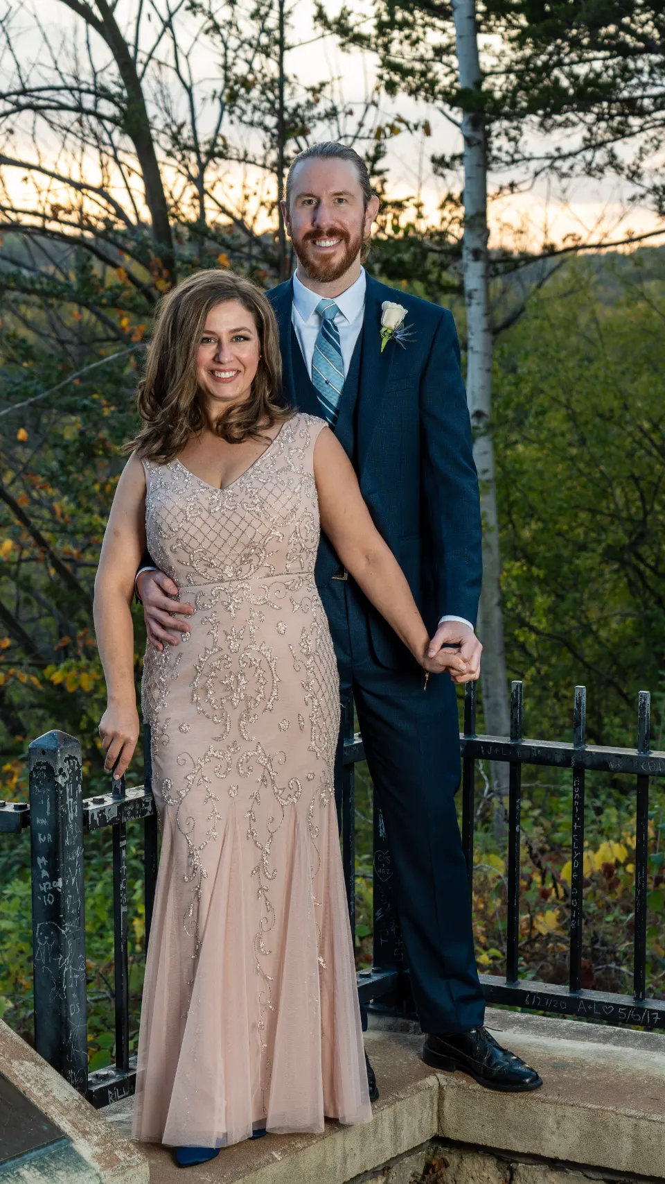 Couple holding hands by a metal railing, in a blue suit and an embellished blush dress.