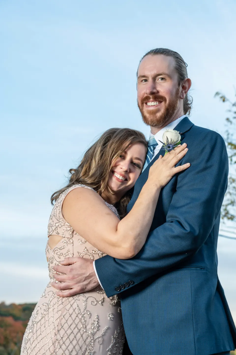Couple embracing beneath a pale blue sky, in a blue suit and an embellished blush dress.