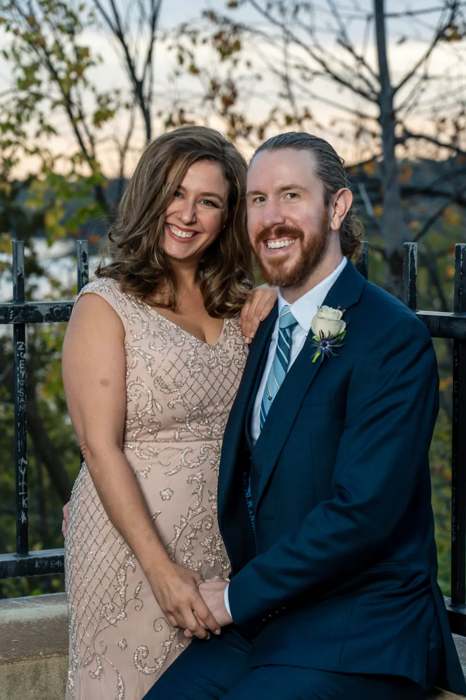 Smiling couple seated by a dark railing, holding hands with trees behind them.