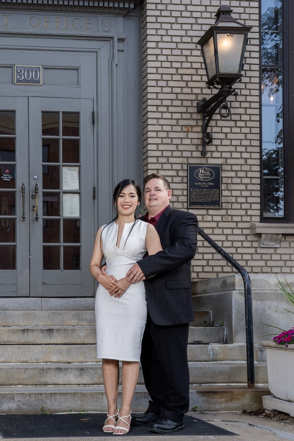 Full-length couple portrait beside brick steps, a gray doorway and a wall lantern.