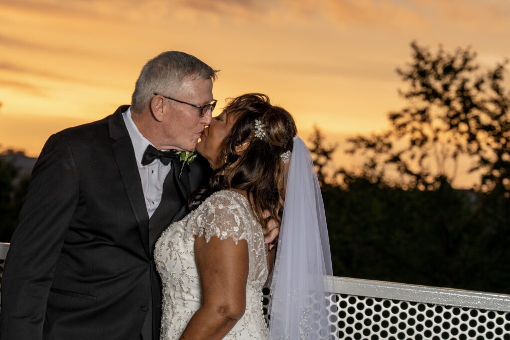 Newlyweds kissing beneath an orange sunset sky.