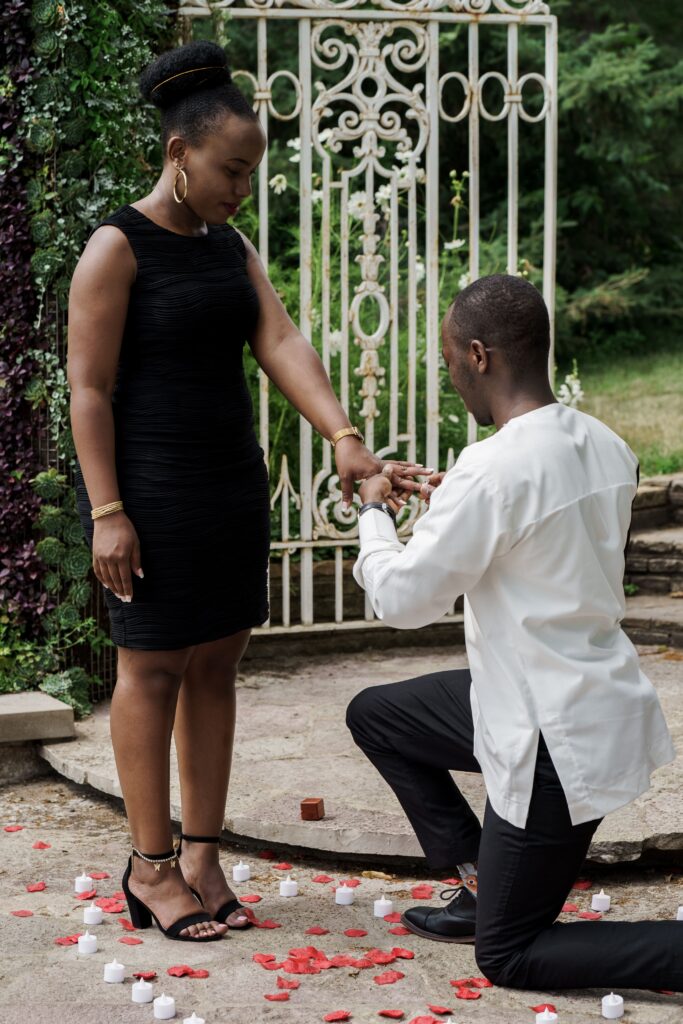 A man kneeling with a ring as his partner stands beside a garden gate.