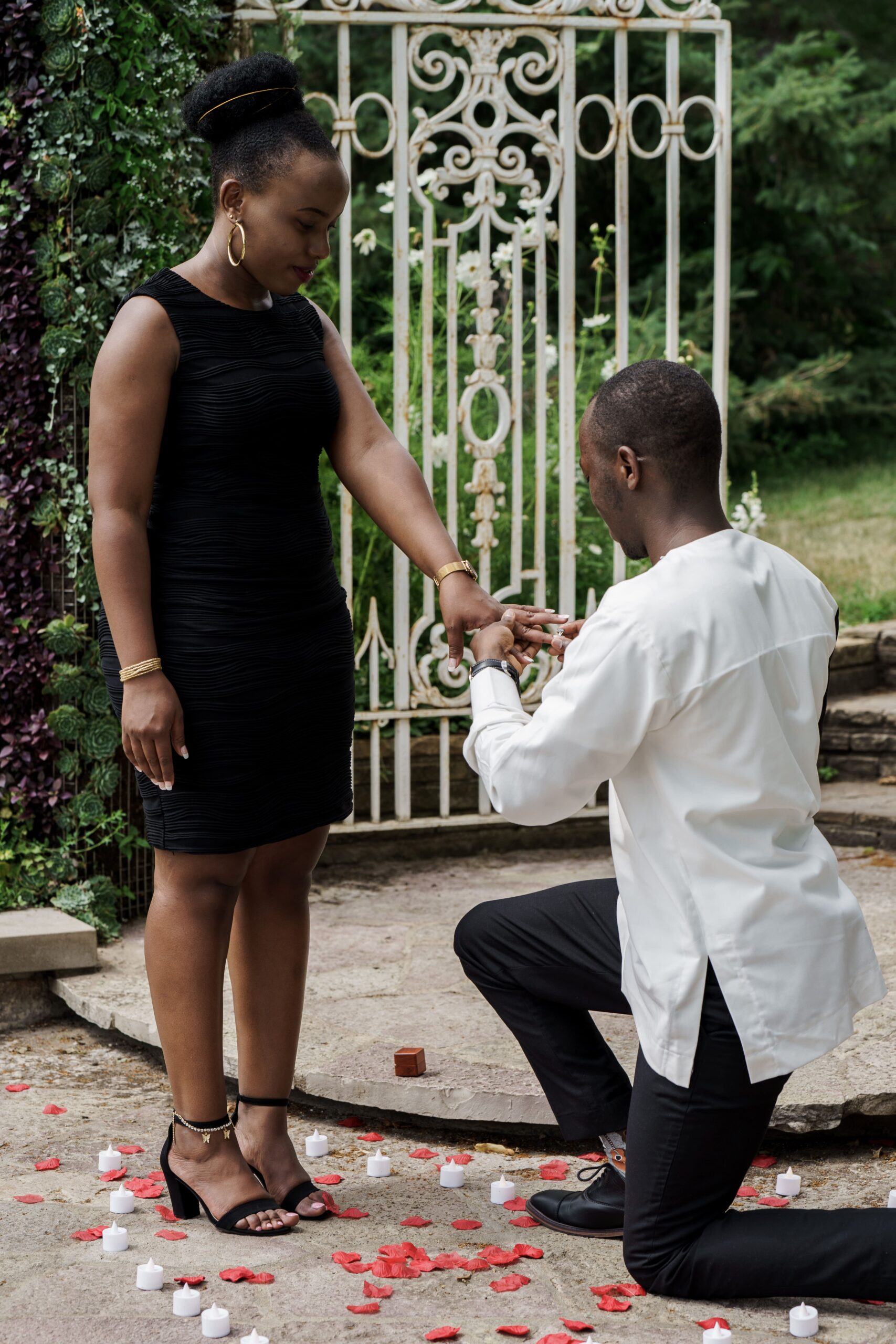 A man kneeling with a ring as his partner stands beside a garden gate.